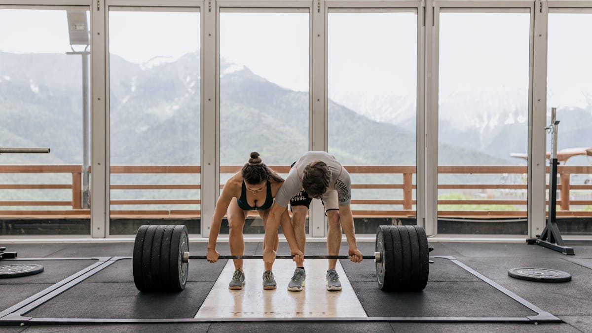 A couple lifting a heavy barbell together in a gym with mountain views.
